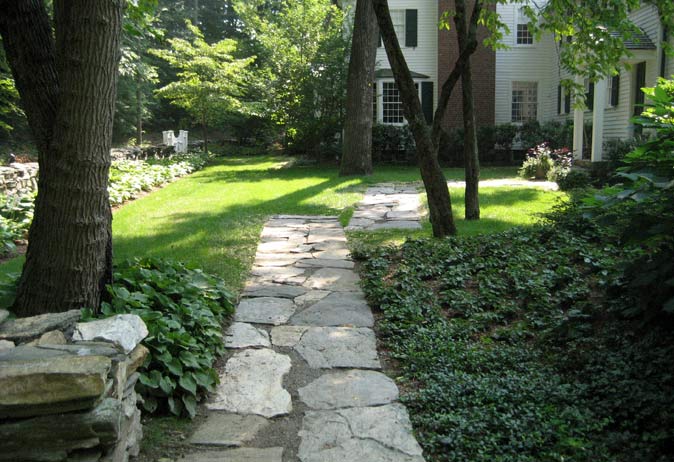 Flagstone pathway through a woodland garden with low groundcover and hostas, leading toward a historic white home.