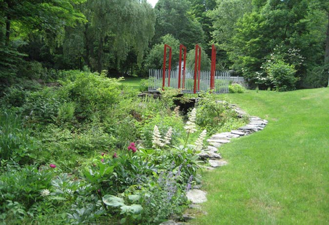 Stone path curving through lush green woodland perennials toward a pond and a modern white and red bridge.
