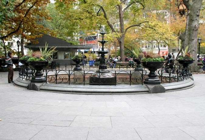 Circular iron fountain with potted plants and a black railing on a bluestone paver platform.
