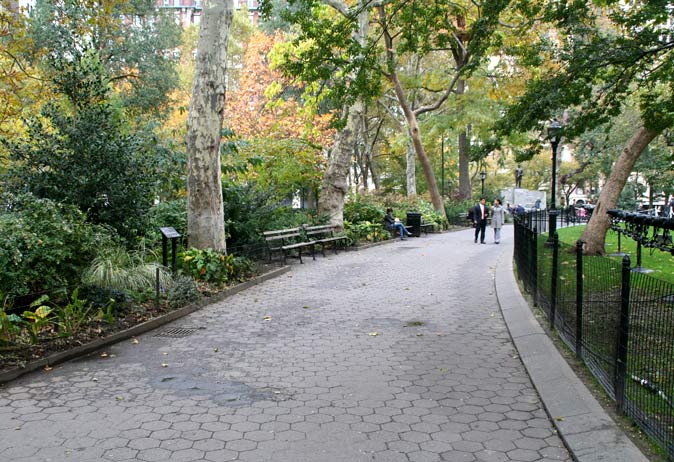Hexagonal-paver path in the park surrounded by lush shrubs, mature trees, a metal fence, and benches.