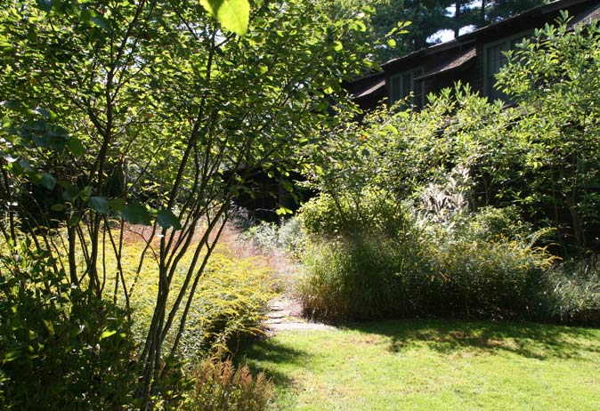 Curved natural stone path through mixed shrubs and tall grasses on a sunny lawn area next to the rustic, shingled home.