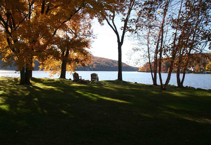 Lakeside lawn seating area with Adirondack chairs and a fire pit, shaded by trees with brilliant fall foliage and lake views.
