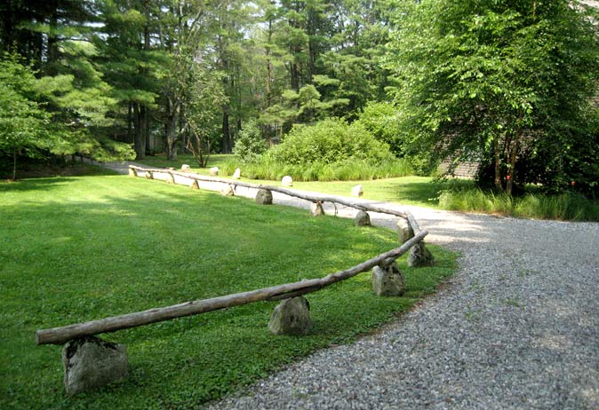 Rustic, curved log railing supported by fieldstones separating a gravel driveway from a lawn.