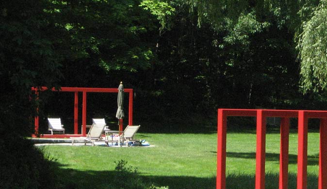 Modern, bright red rectangular arbor structures framing a lounge area on a green lawn backed by dense woods.