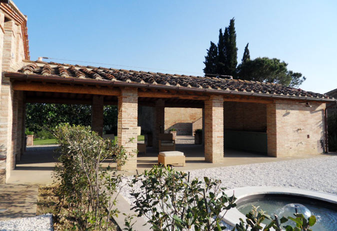 Covered outdoor living portico with terracotta roof and brick columns, offering shade over a gravel courtyard.