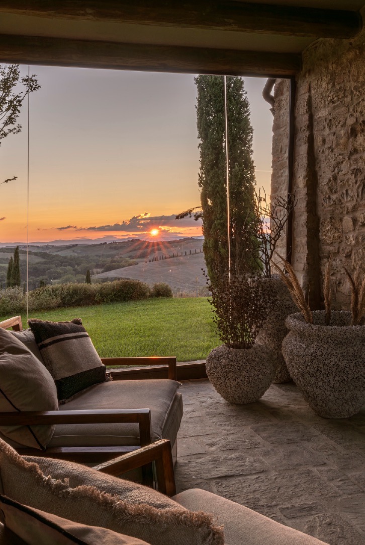 Stone porch view of a Tuscan sunset through glass, with rustic lounge chairs and large textured planters.