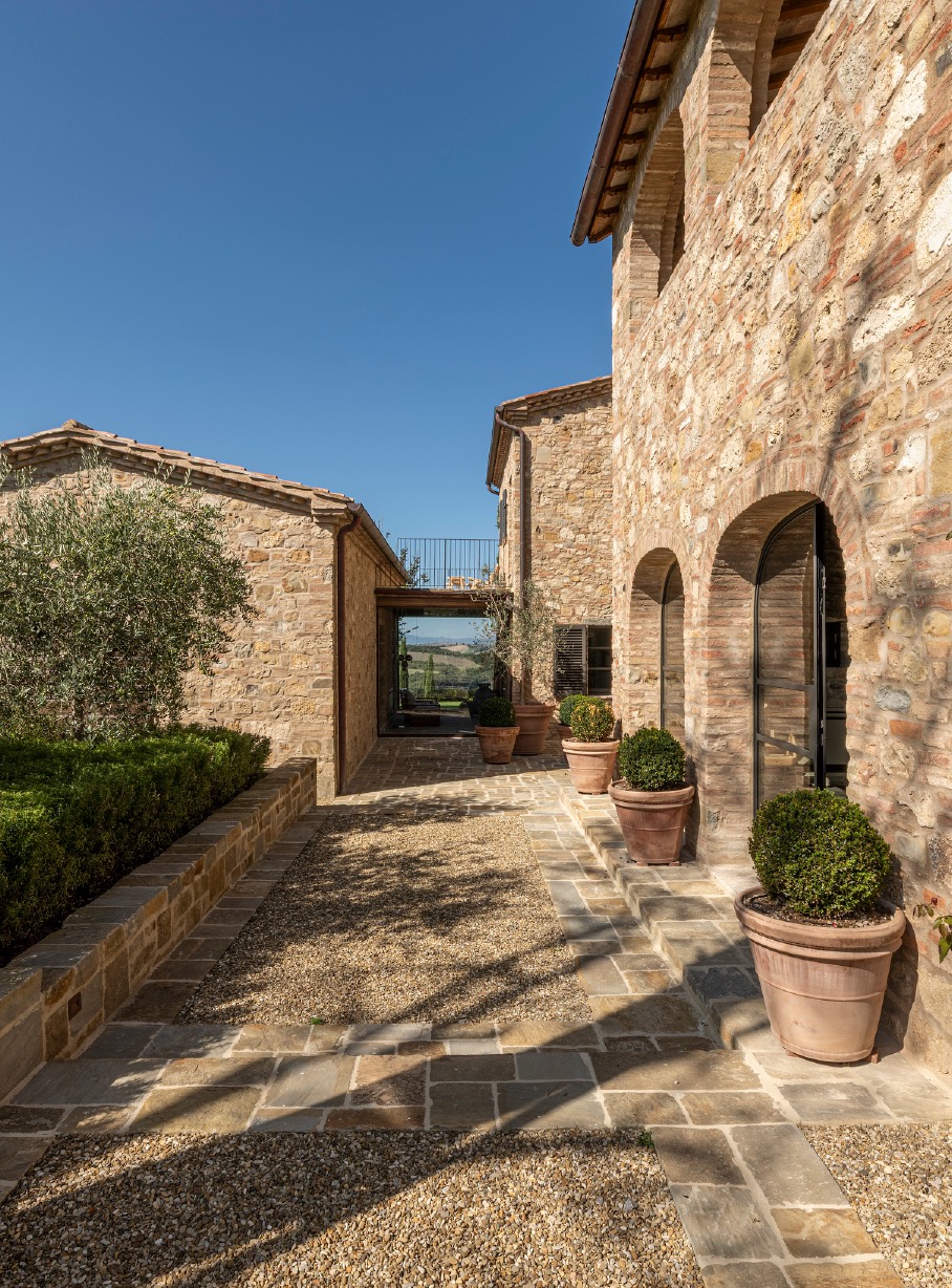Stone walkway with potted boxwood globes between rustic buildings, framing a view of distant hills.