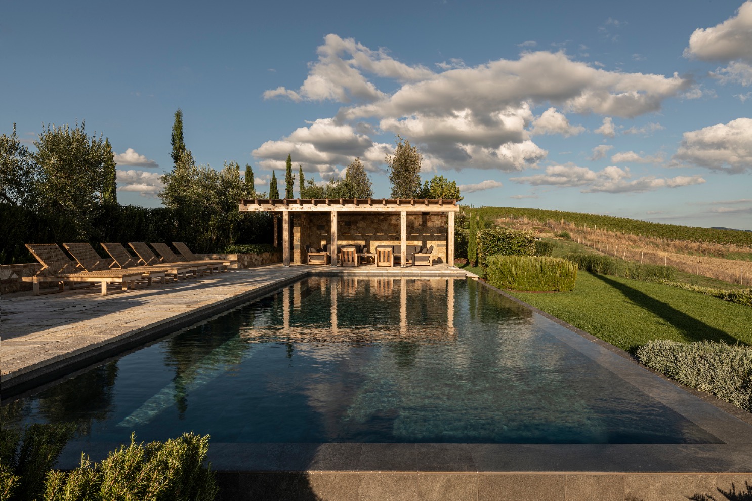 Infinity pool reflecting a cloudy sky, featuring a wooden pergola lounge and Mediterranean plantings.