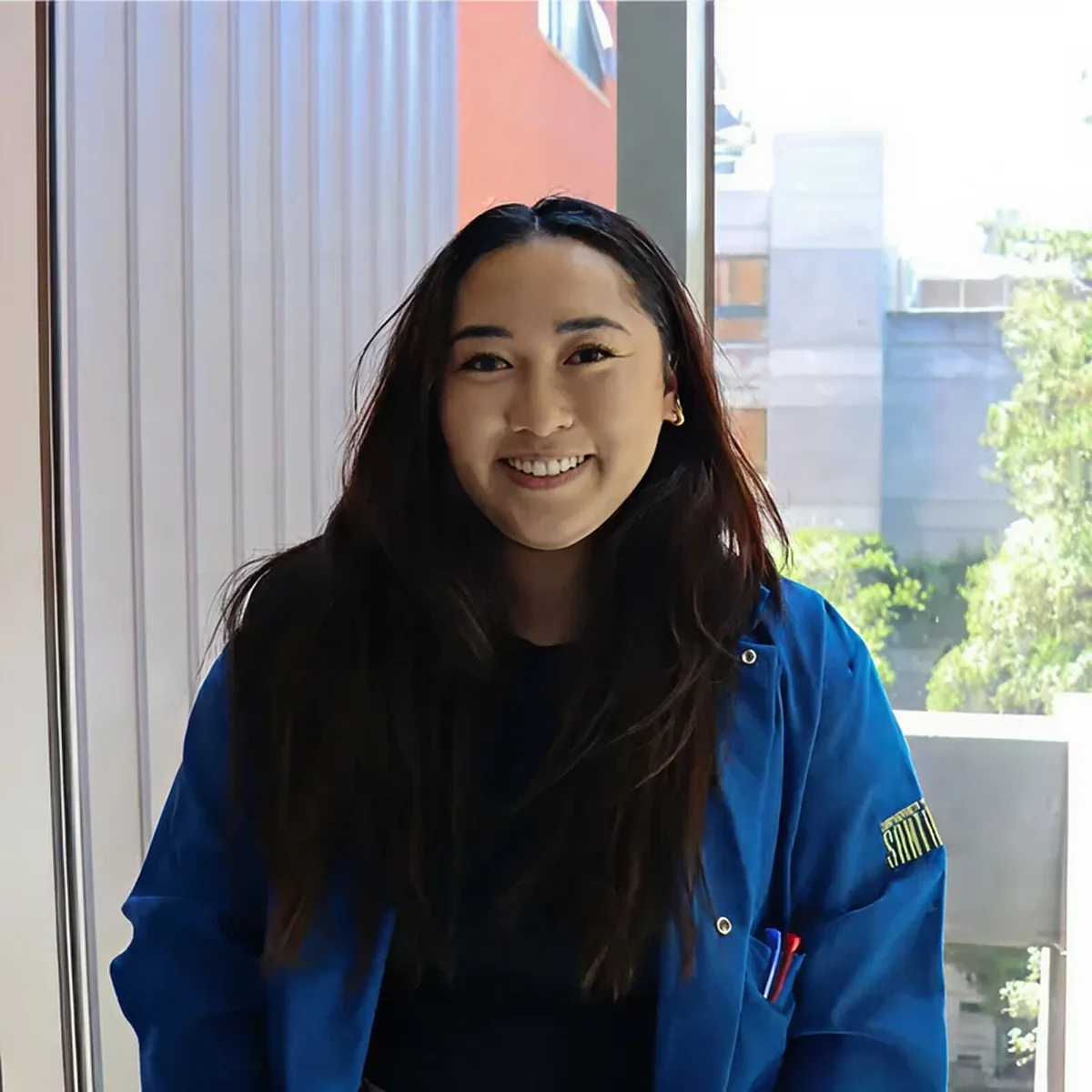 Smiling woman with long dark hair wearing a blue jacket standing indoors near a window with greenery outside.