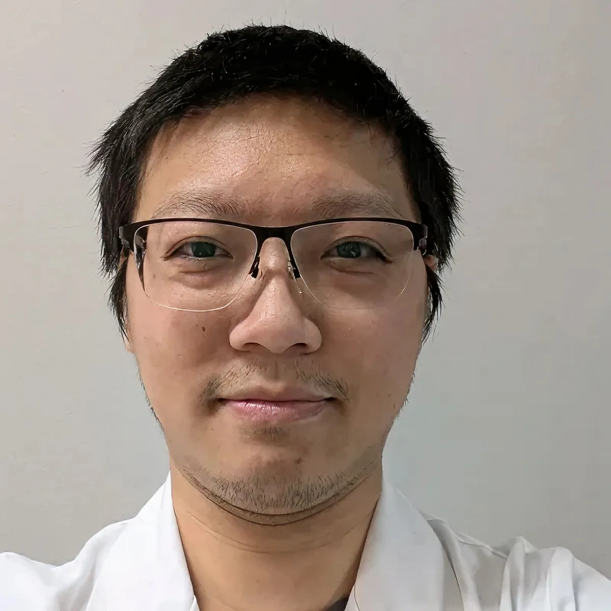 Close-up portrait of a man with short black hair and glasses wearing a white shirt, smiling slightly against a plain light background.