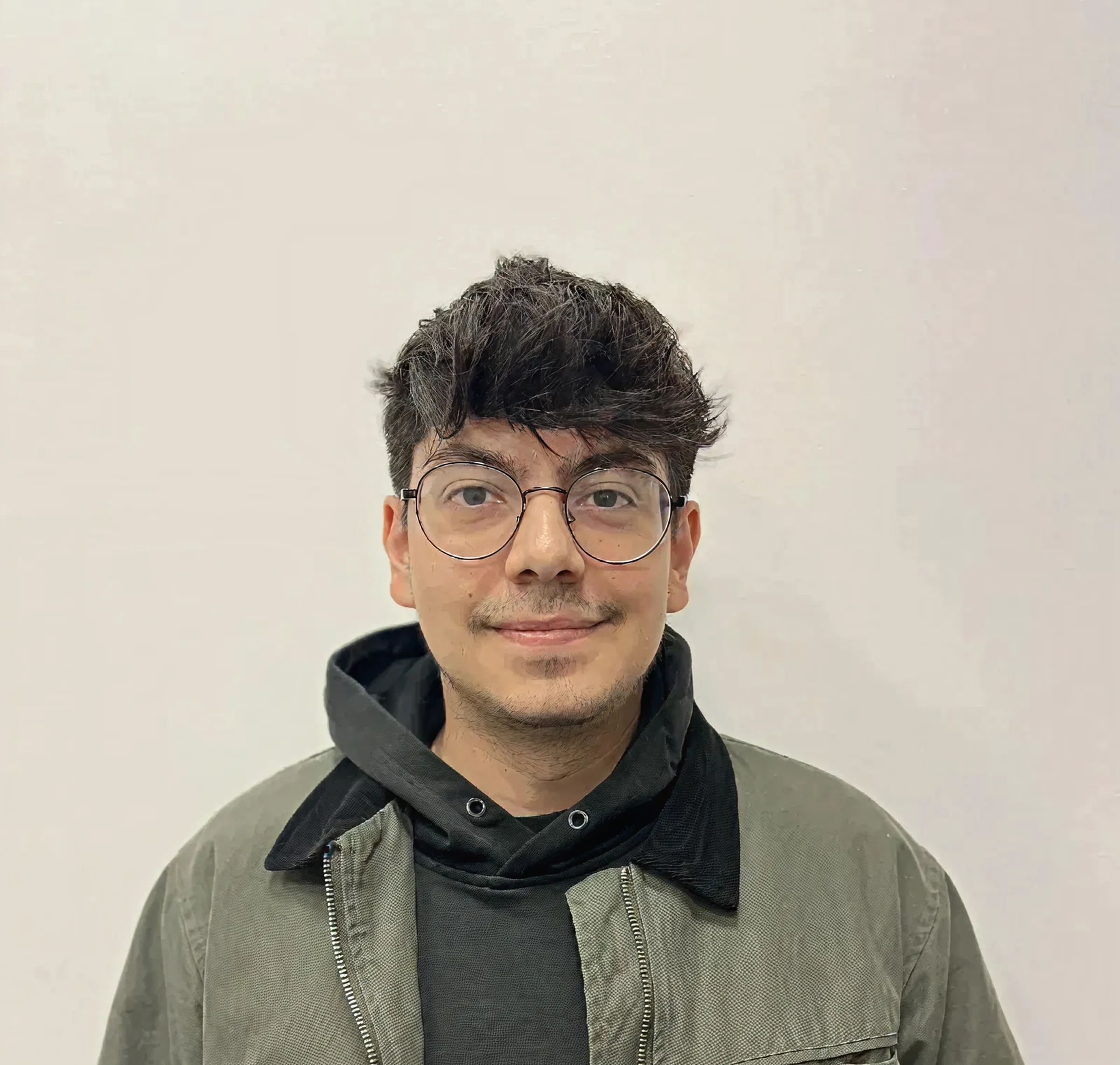 Young man with dark hair and round glasses wearing a green jacket and black hoodie, smiling at the camera against a plain background.