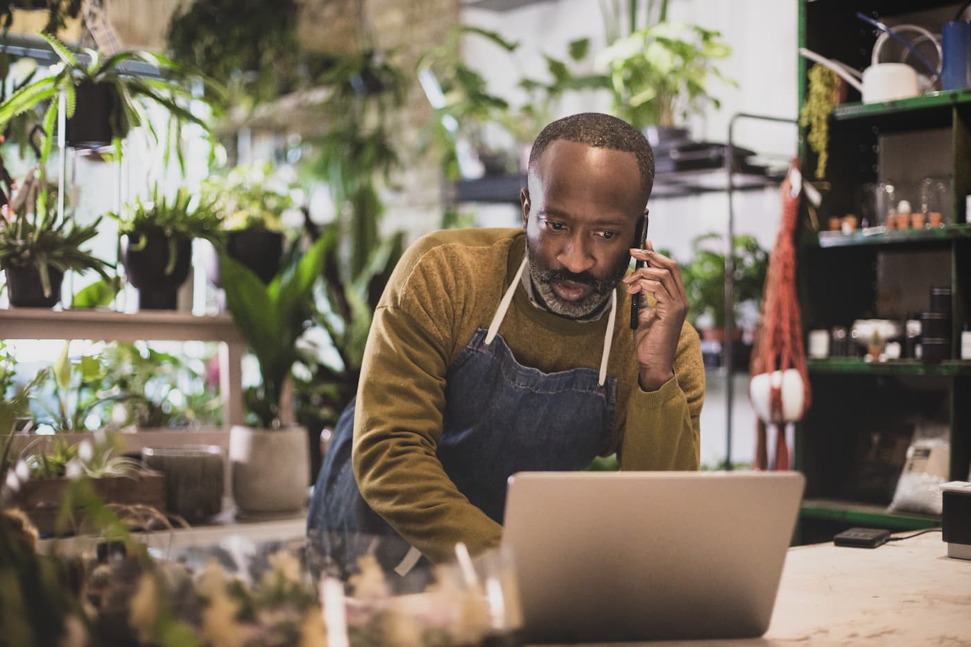 Man wearing apron using a laptop and talking on the phone in a plant shop.
