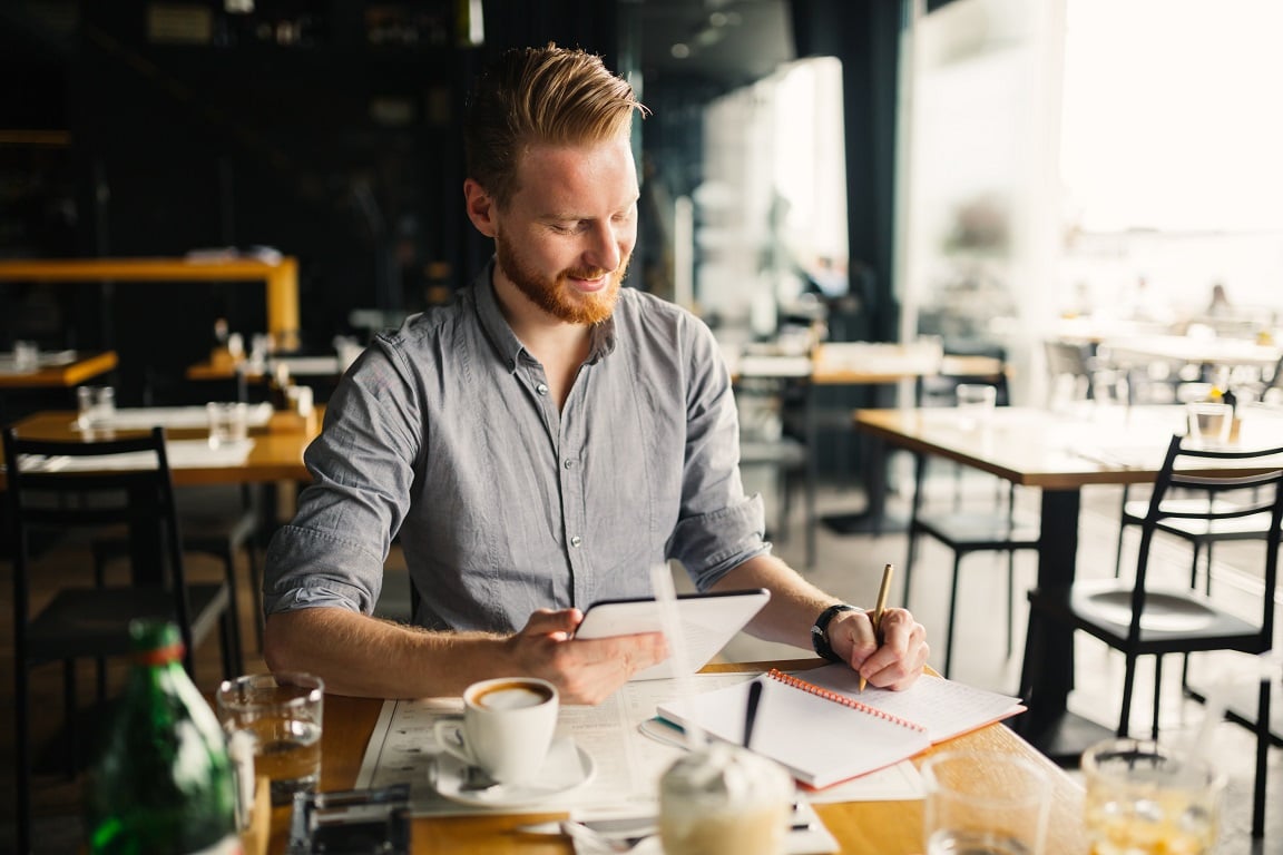 A man with red hair and a beard sitting at a café table, holding a tablet and writing in a notebook, with a cup of coffee and drinks nearby.
