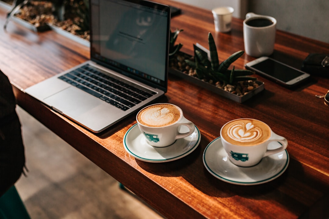 Two cappuccinos with latte art on saucers next to a laptop on a wooden table with potted plants and a smartphone.