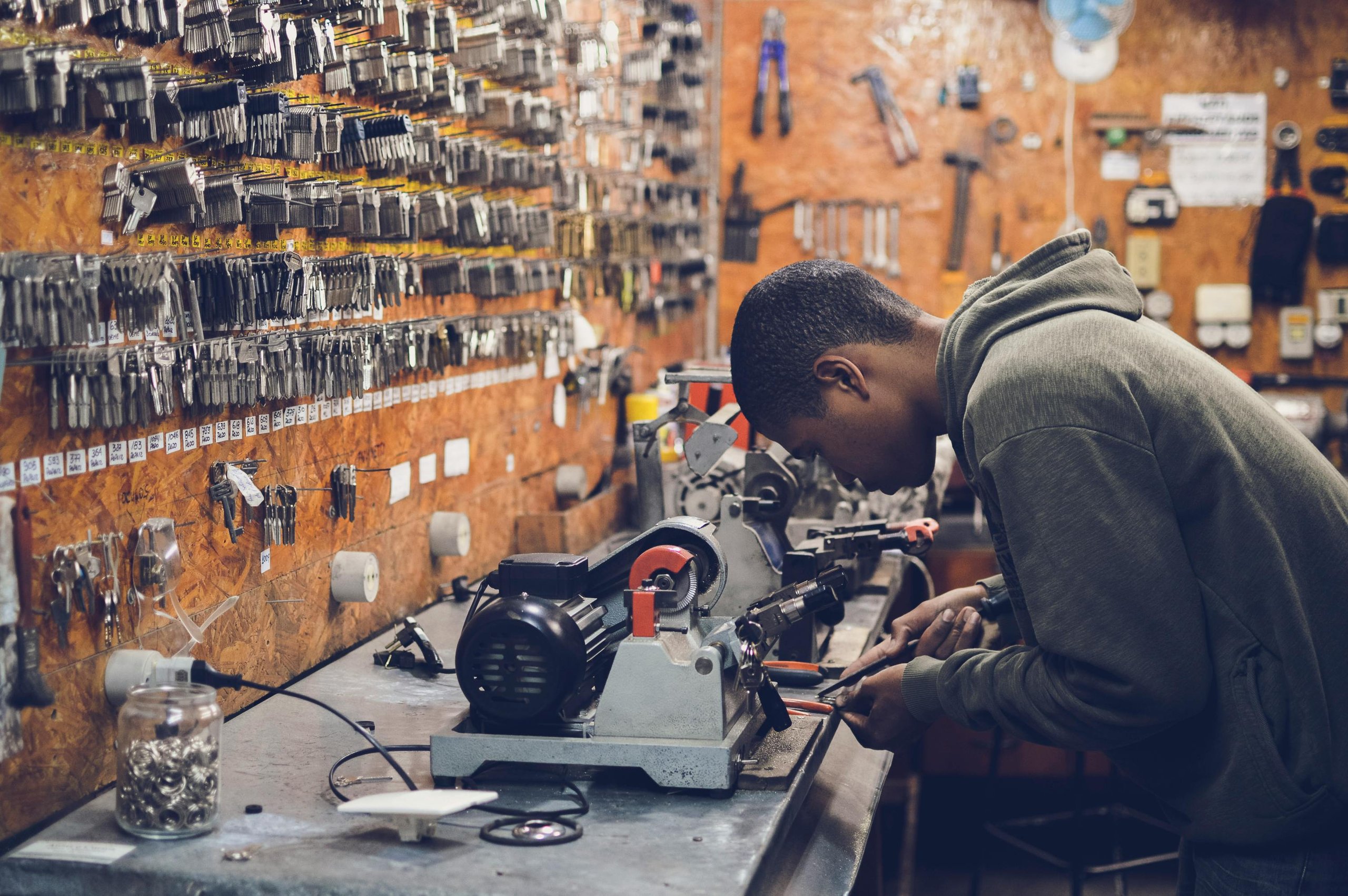 A young man in a gray hoodie working with tools at a key cutting machine in a workshop filled with hanging keys on wooden walls.