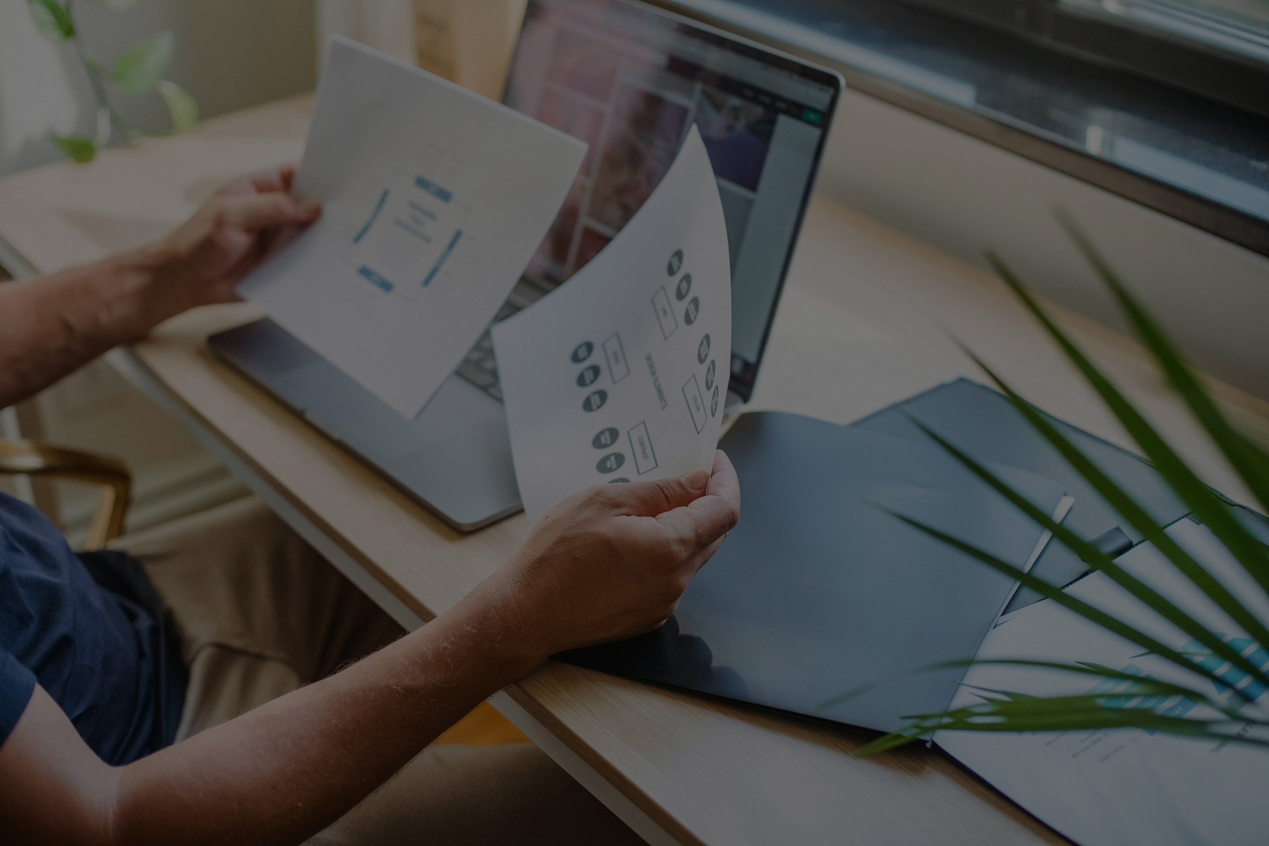 Person holding two paper documents with workflow diagrams over a laptop on a wooden desk.