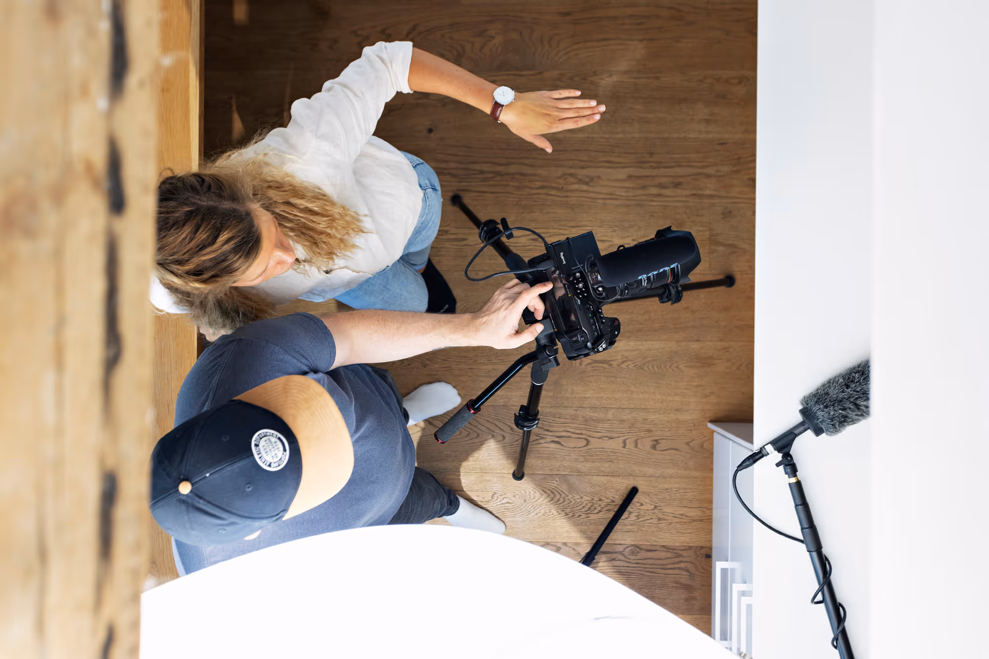 To personer ser på en skjerm på et kamera montert på et stativ på et tregulv med en profesjonell lydmikrofon til høyre.