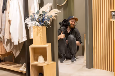 En mann med caps sitter på gulvet i et prøve-rom mens han holder et videokamera og gir tommel opp.