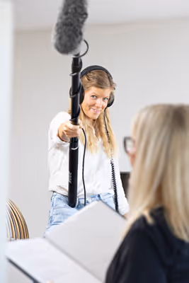 Kvinne med hodetelefoner holder en boom-mikrofon mot en person som leser fra en bok.