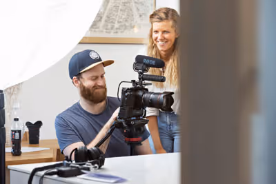 Mann justerer et speilreflekskamera med mikrofon mens en kvinne ser på og smiler i et lyst innendørs studio.