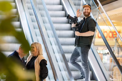 Smilende mann med kamera gir tommel opp foran rulletrapp i kjøpesenter.
