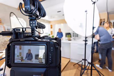 Et profesjonelt kamera som filmer en kvinne i et moderne kjøkkenstudio med lysutstyr og en fotograf i bakgrunnen.