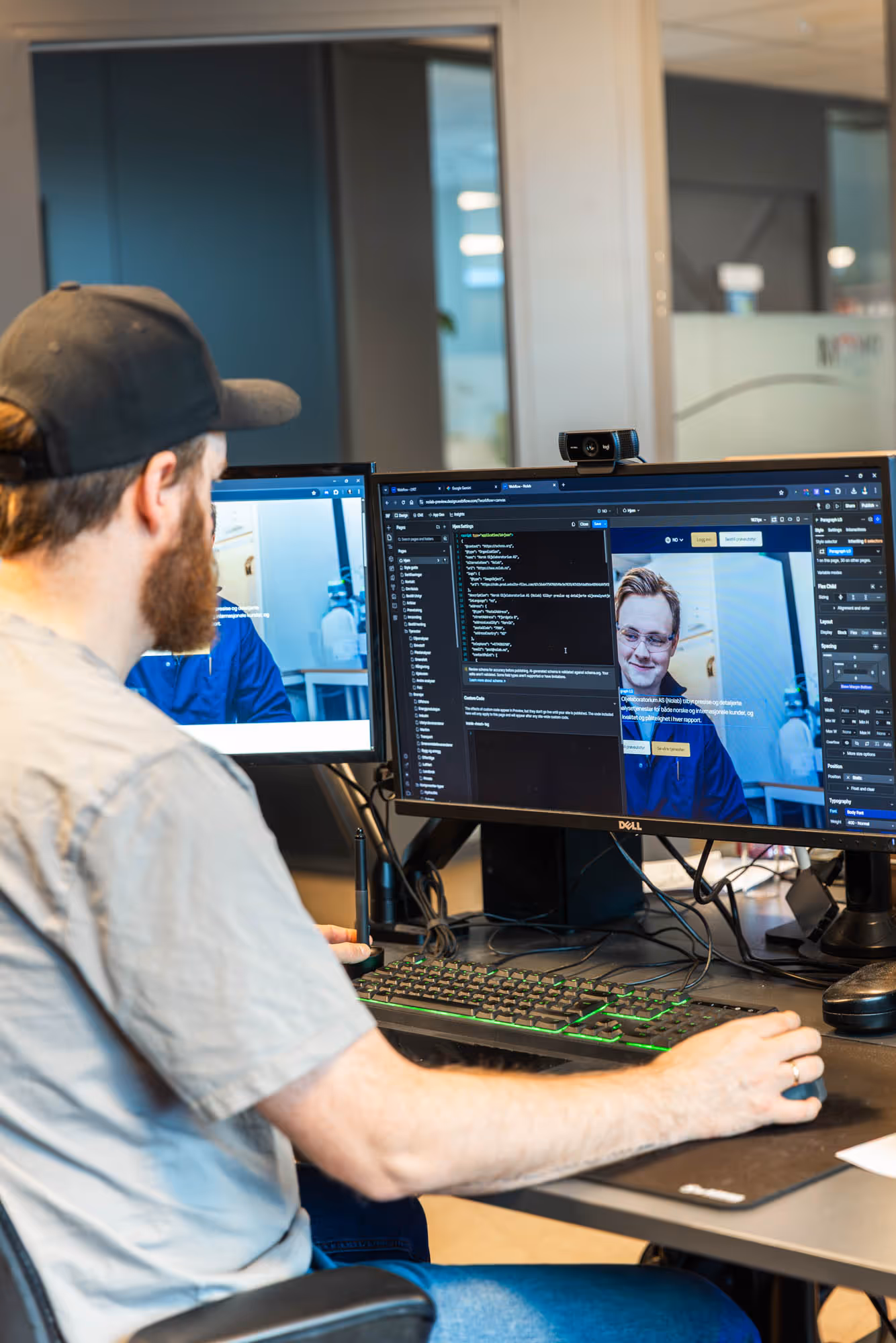 En mann med baseballcap sitter ved et skrivebord og arbeider på en datamaskin med to skjermer som viser programmeringskode og en videosamtale.