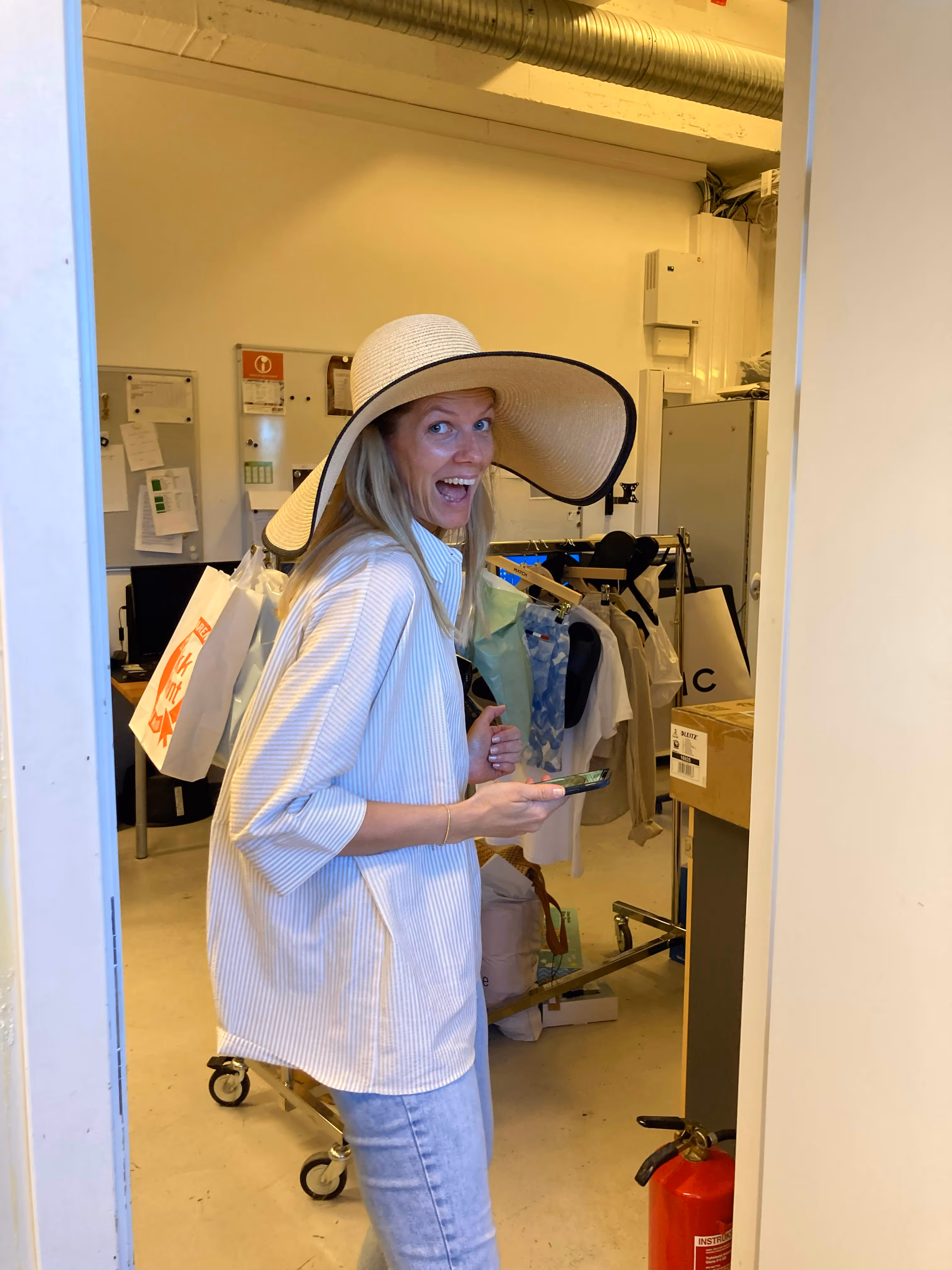 Kvinne i stor hvit hatt, stripete skjorte og jeans smiler bredt mens hun holder en mobiltelefon i et rom med klær og esker.