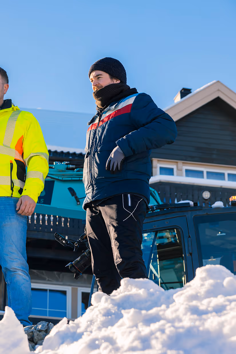 Mann i vinterjakke og lue står ved snø med et kamera i hånden foran et hus og en blå maskin.