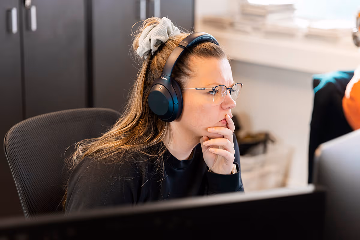 Kvinne med briller og svarte Sony-hodetelefoner som ser konsentrert på en dataskjerm i et kontormiljø.