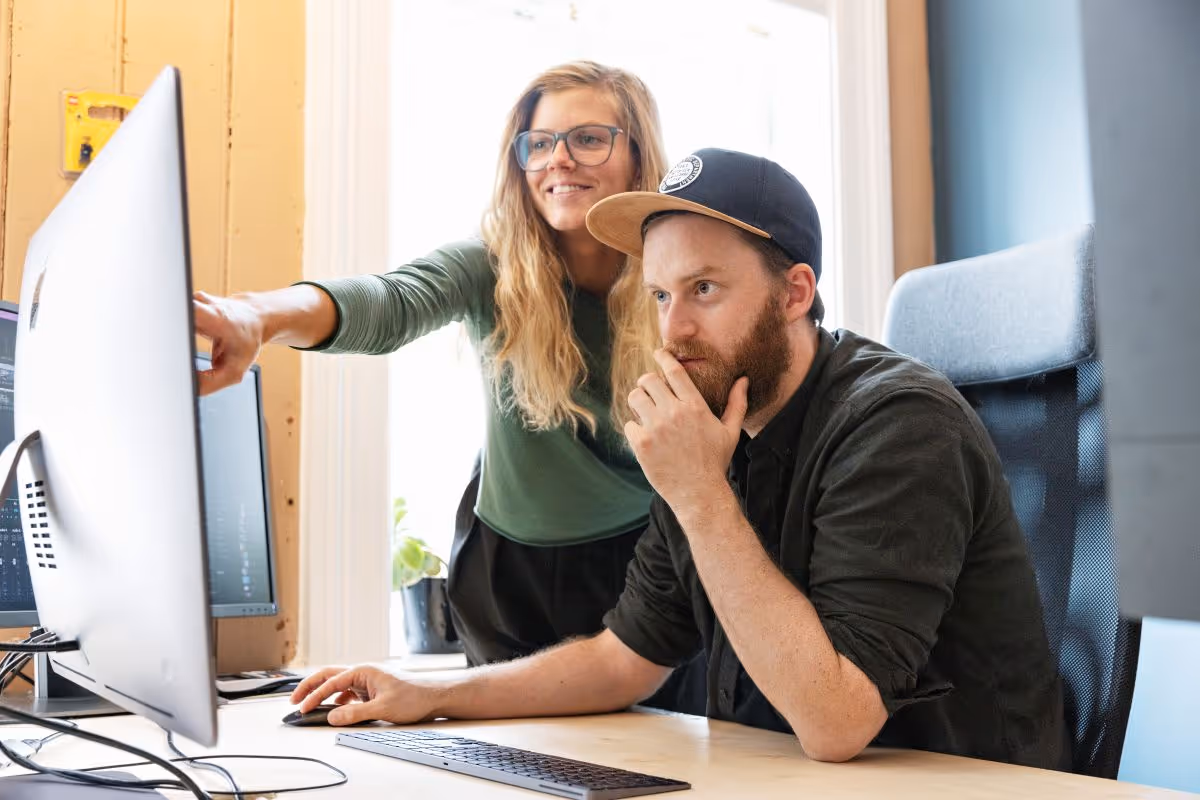 To kolleger diskuterer noe på en dataskjerm ved et skrivebord i et lyst kontor.