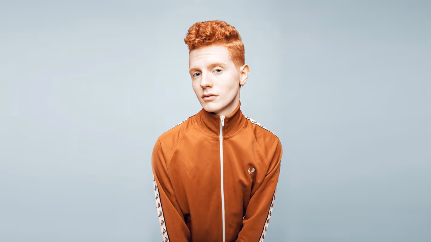 Ung mann med rødt krøllete hår iført oransje zip-up treningsjakke mot lys bakgrunn.