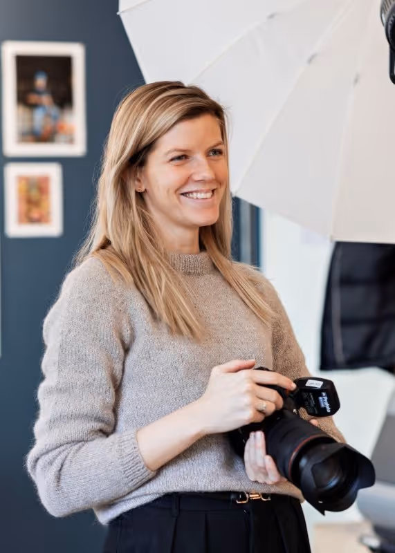 Kvinne med blondt hår smilende og holder et profesjonelt kamera i et fotostudio.