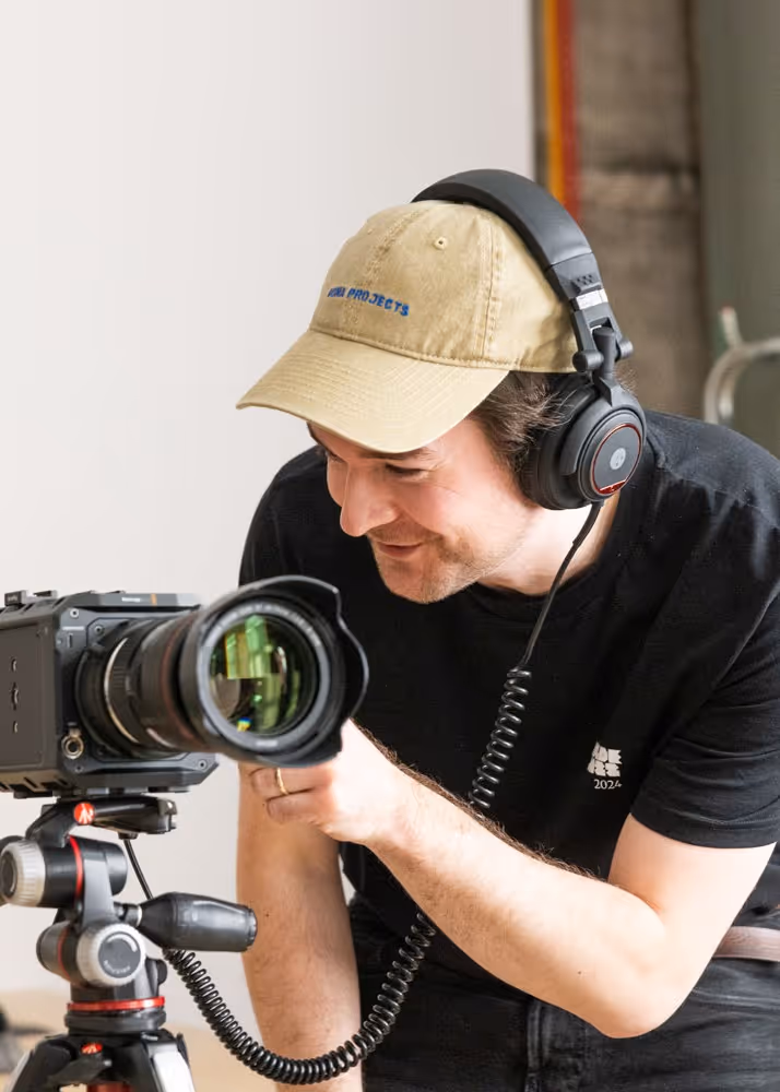 Mann med beige caps og sorte hodetelefoner som ser gjennom et kamera på et stativ.