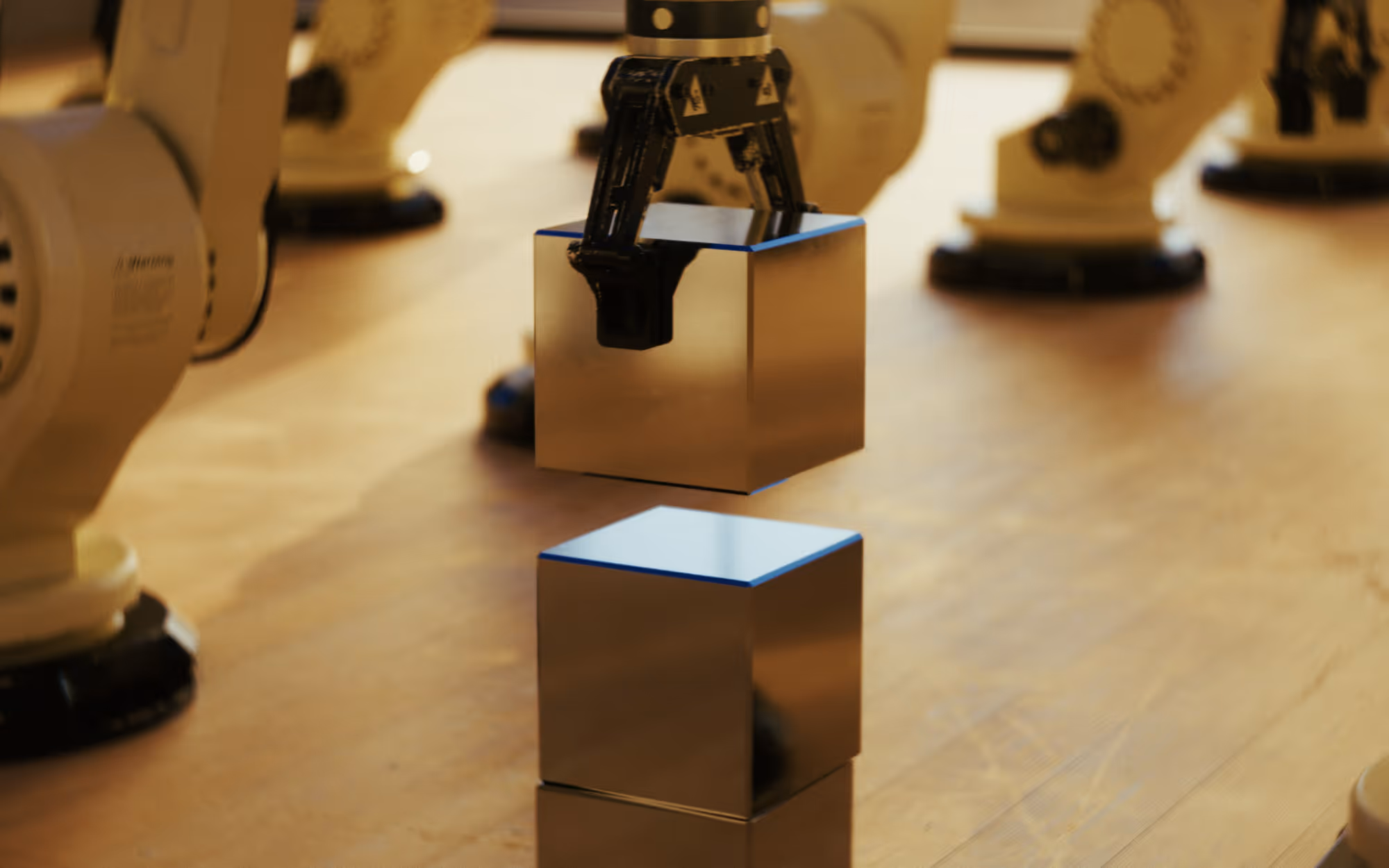 Robot arm lifting a reflective silver cube above another identical cube on a wooden surface.