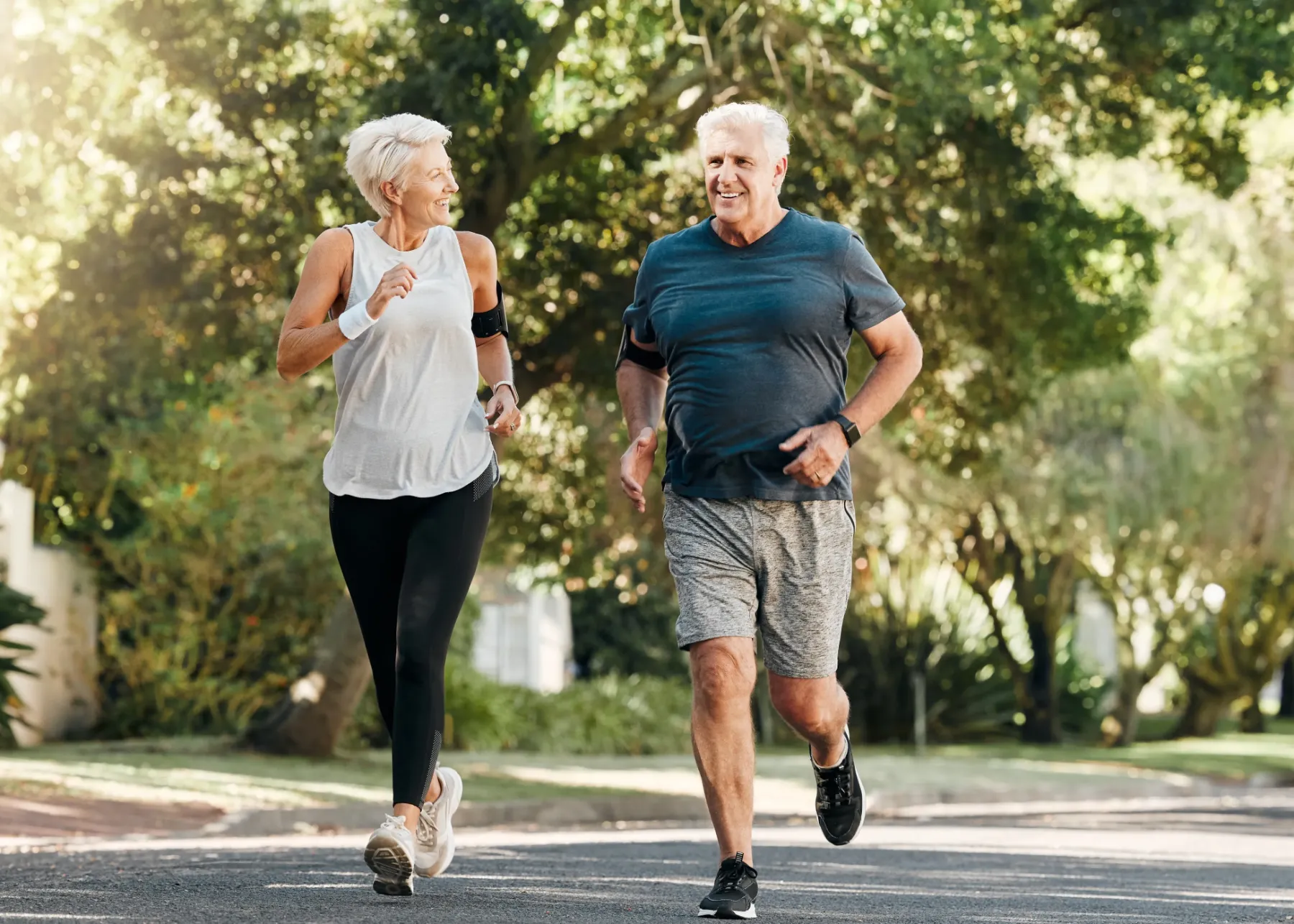Smiling senior man and woman jogging outdoors on a sunny day along a tree-lined street.