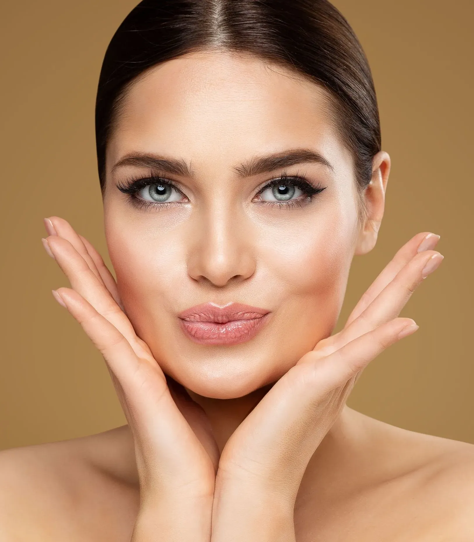 Close-up of a woman with smooth skin, blue eyes, and brown hair holding her face with both hands.