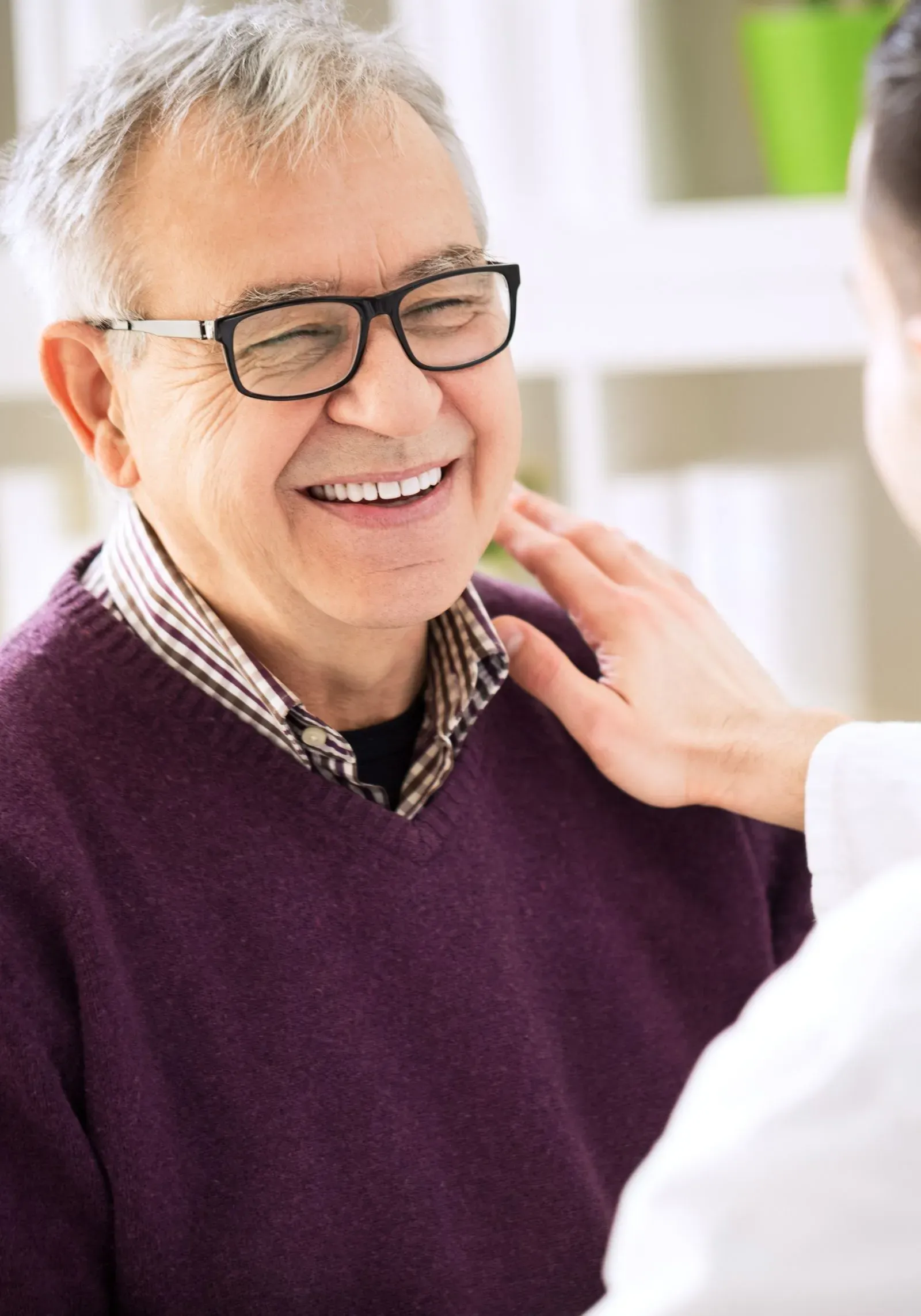 Smiling elderly man wearing glasses and a purple sweater being gently touched on the shoulder by another person.
