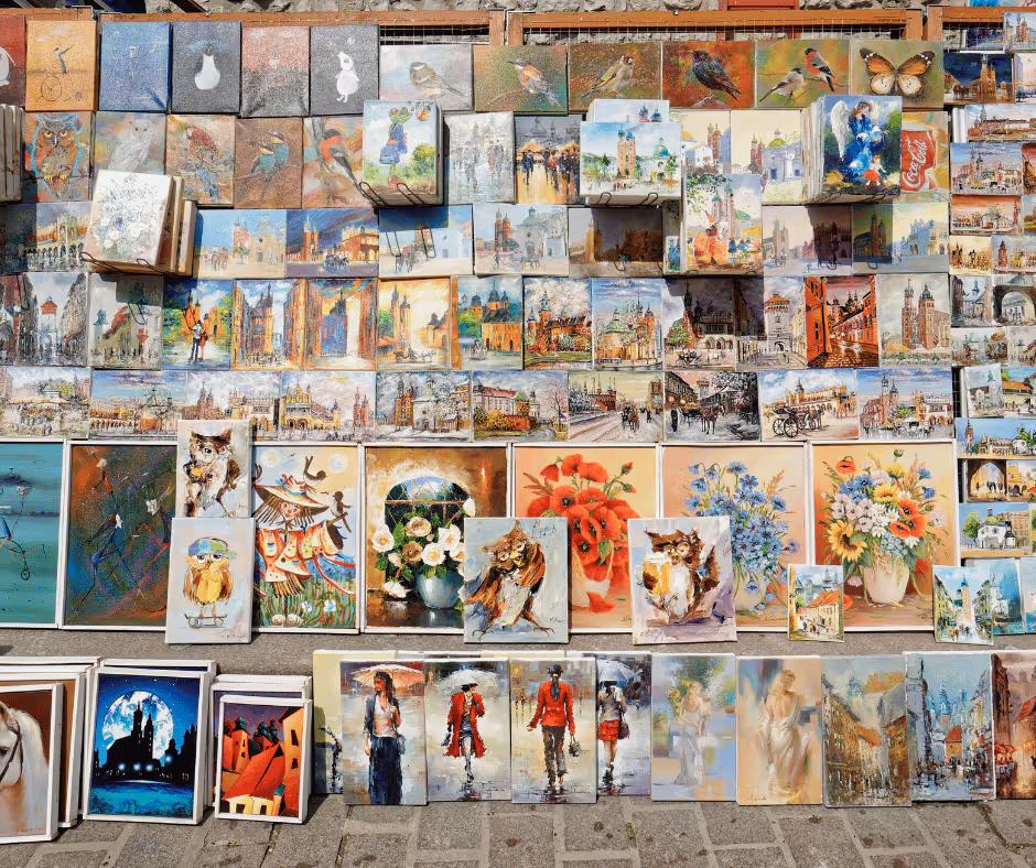 Wall display of colorful paintings featuring owls, flowers, cityscapes, and street scenes arranged outdoors on a sunny day.