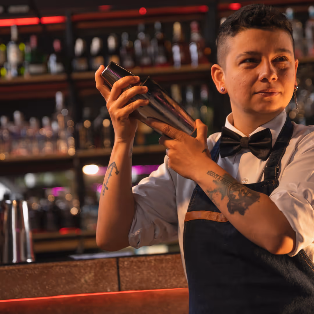 Bartender in white shirt, black bow tie, and apron shaking a cocktail shaker behind a bar.