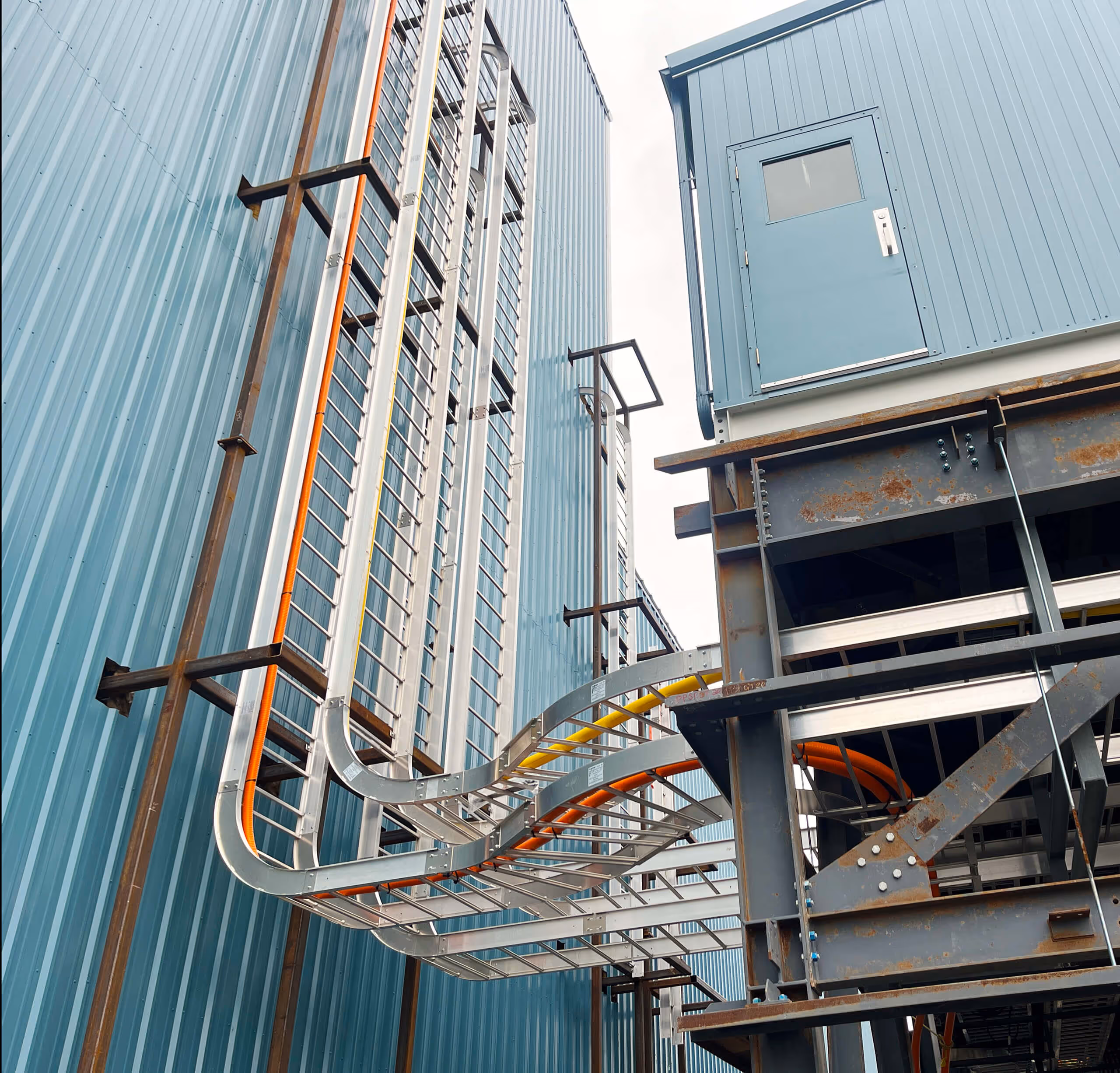 Exterior view of blue industrial buildings with exposed metal cable trays carrying colored wiring between the structures