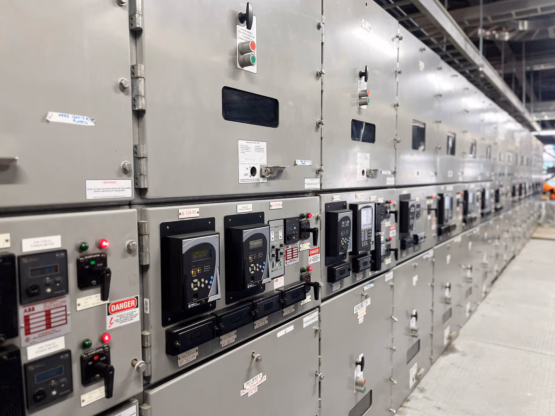 Long row of industrial electrical control panels with meters, switches, and warning labels in a power distribution room.