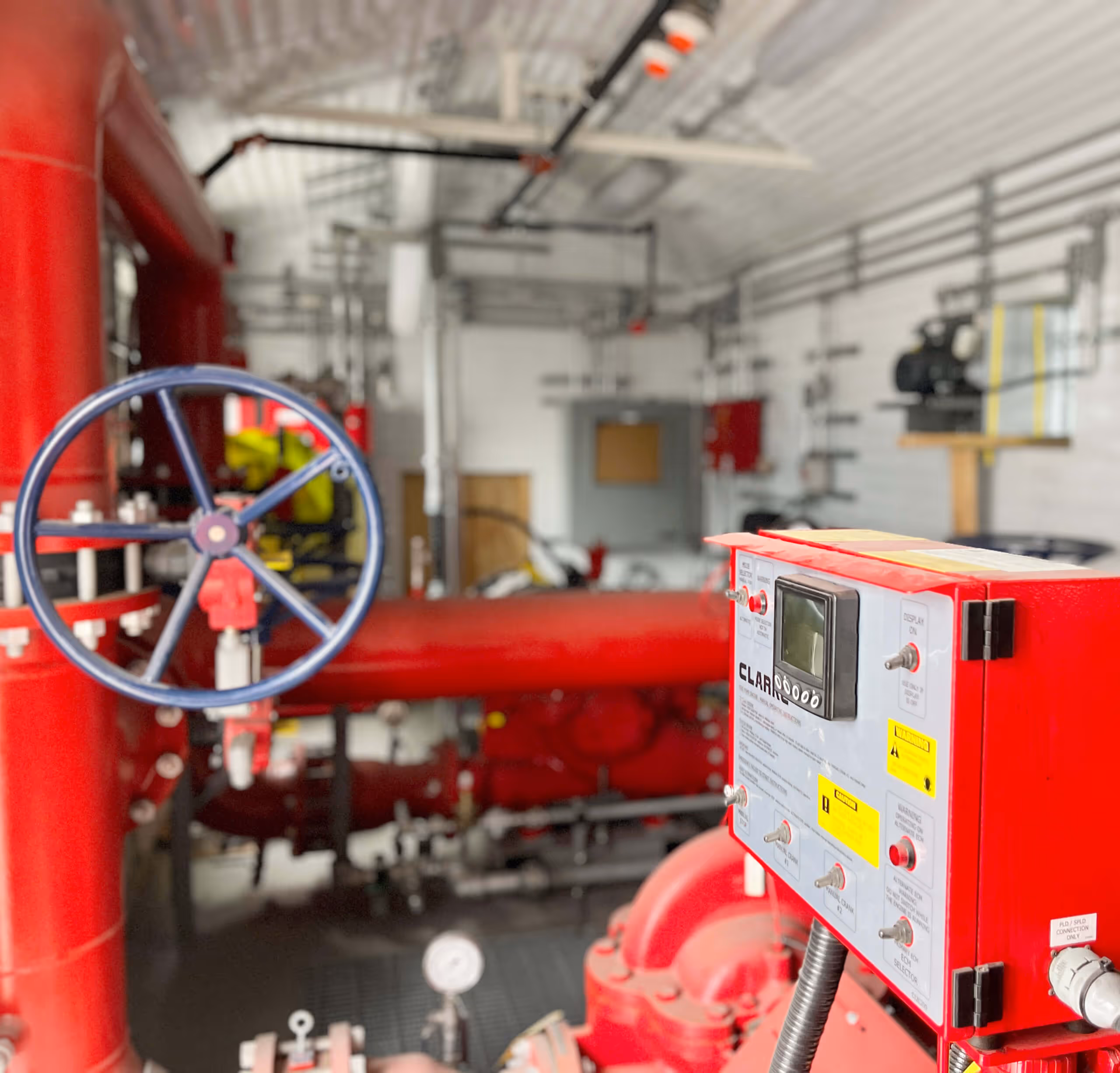 Red fire pump control panel in a sprinkler room with large red pipes and a blue manual valve wheel