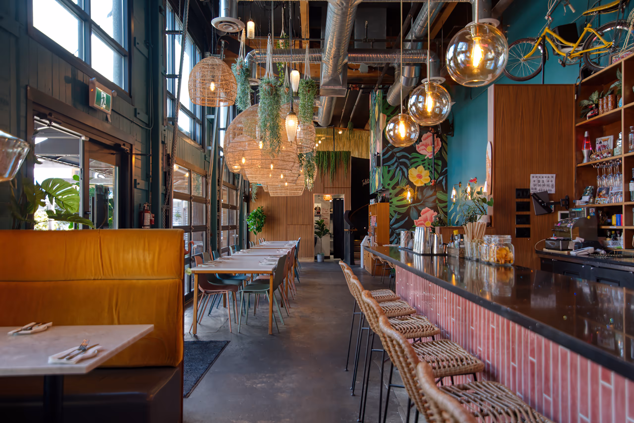 Restaurant interior with wicker pendant lights, floral mural, and a long bar with woven stools.