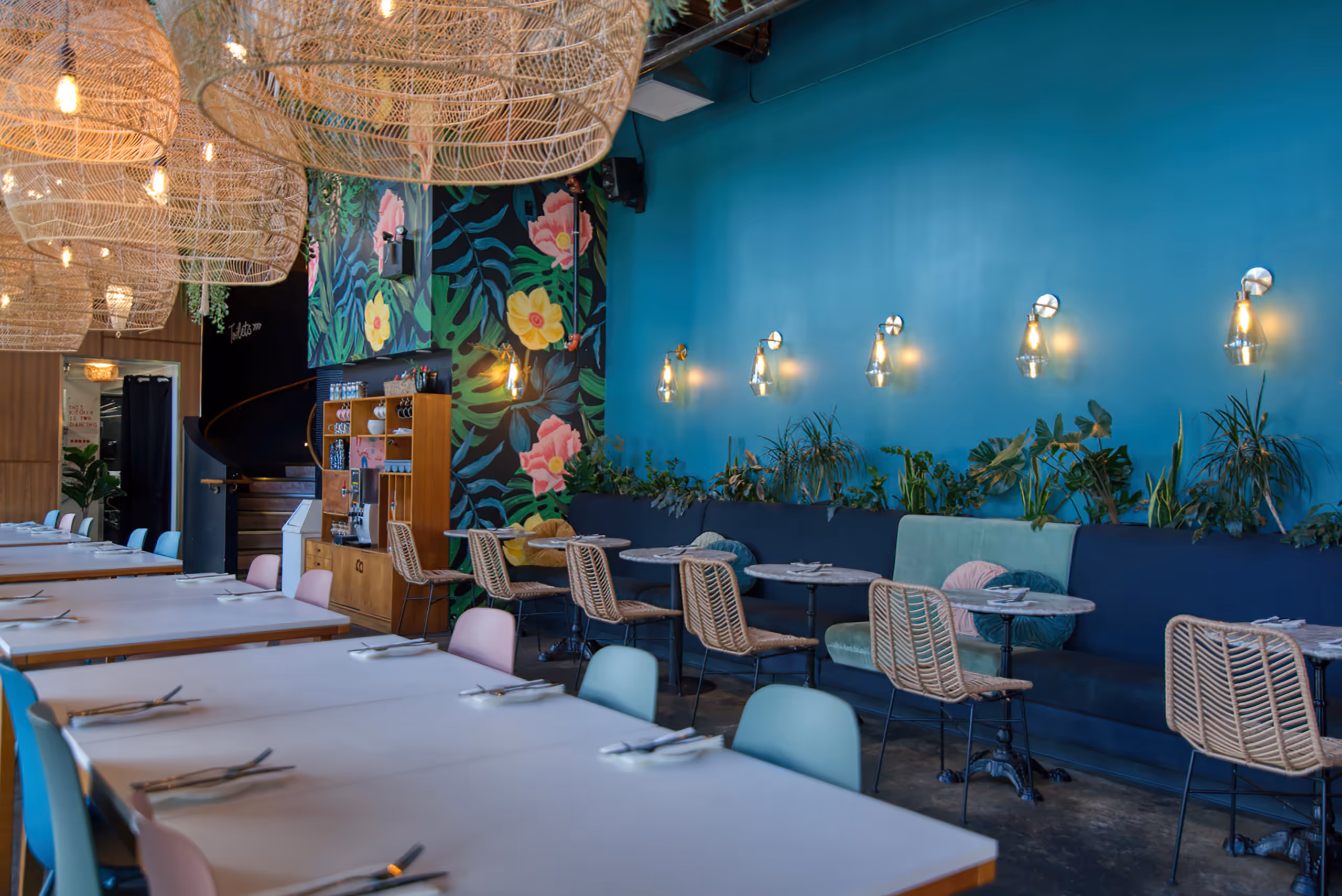 Colorful restaurant interior with teal walls, floral mural, and wicker light fixtures.