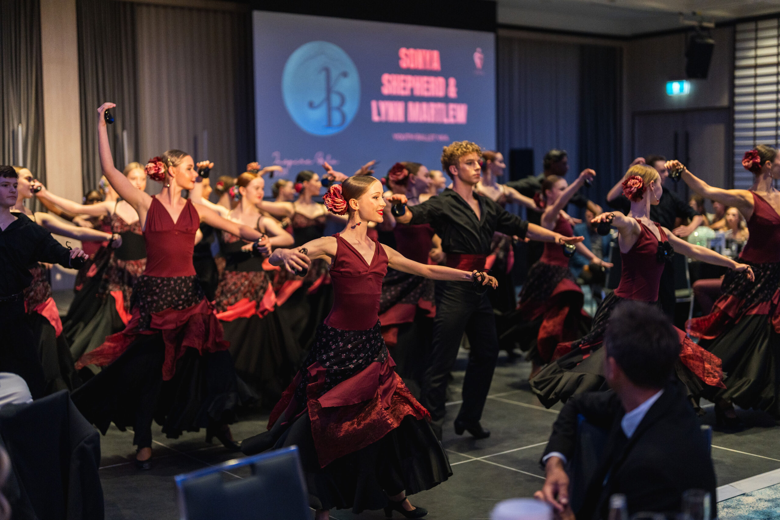 Group of dancers in coordinated red and black costumes performing a flamenco dance indoors with audience watching.