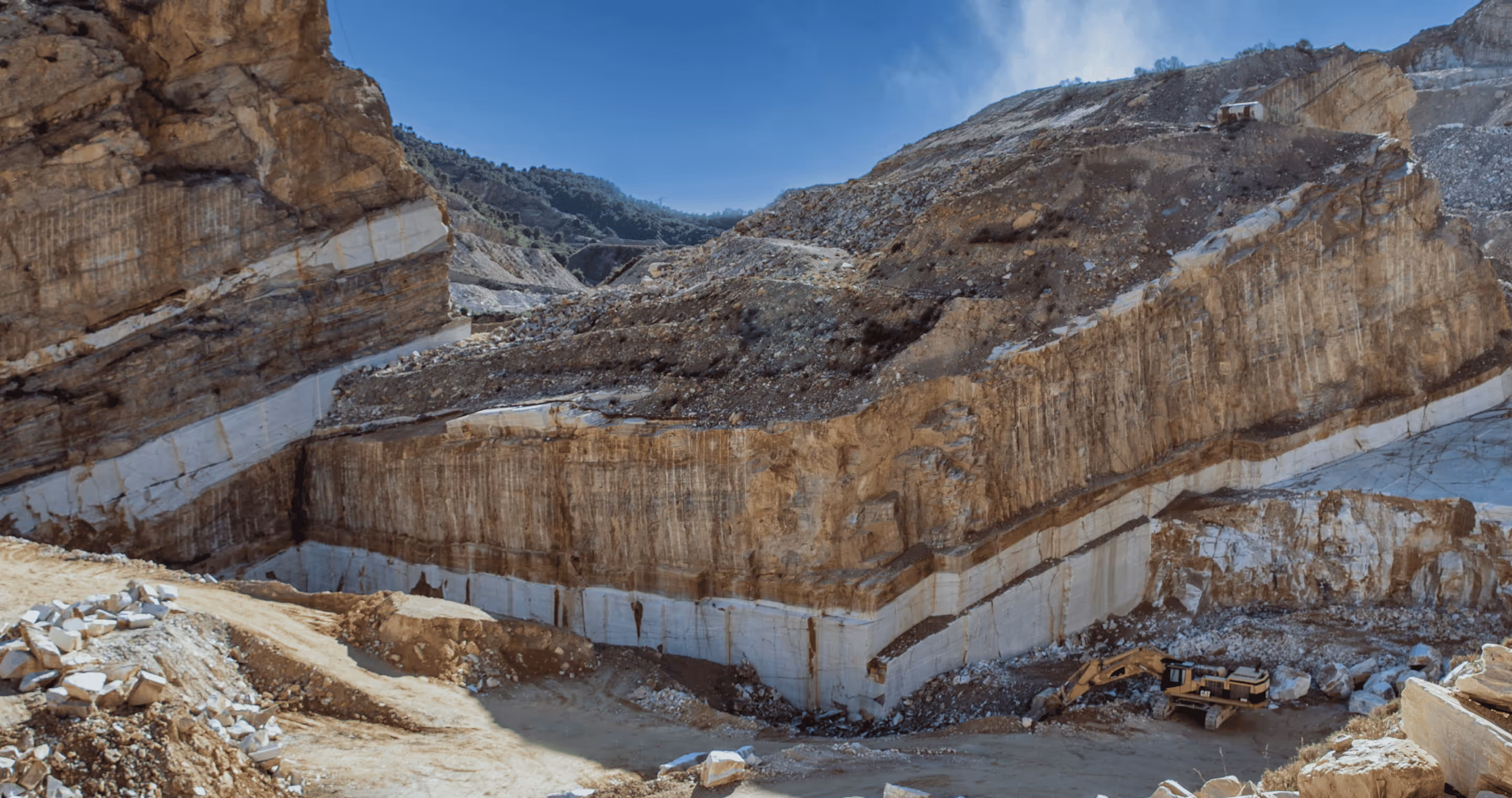 Open quarry with layered rock formations and an excavator at the bottom under a clear blue sky.