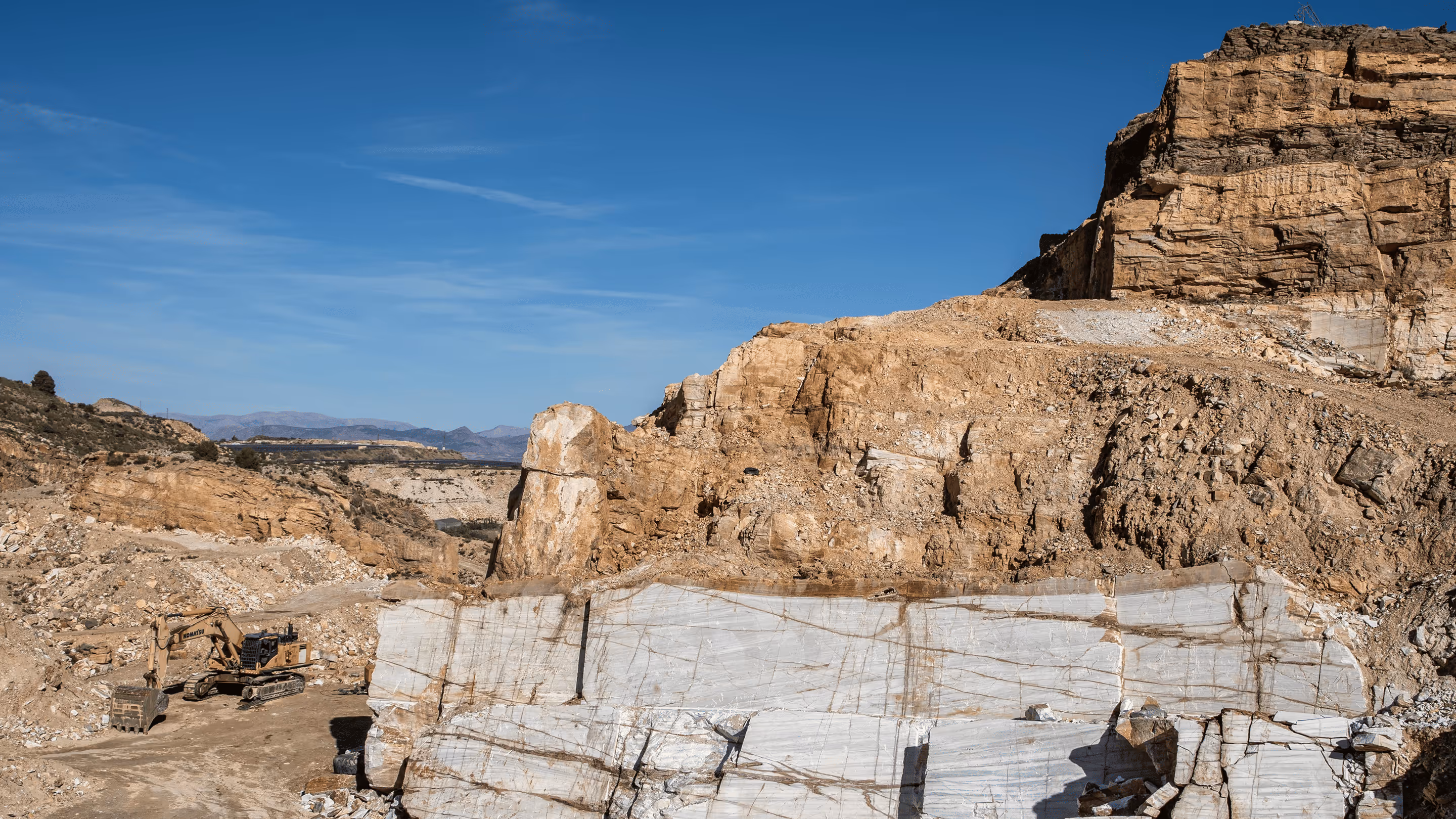Marble quarry with large white stone blocks and an excavator against a clear blue sky.