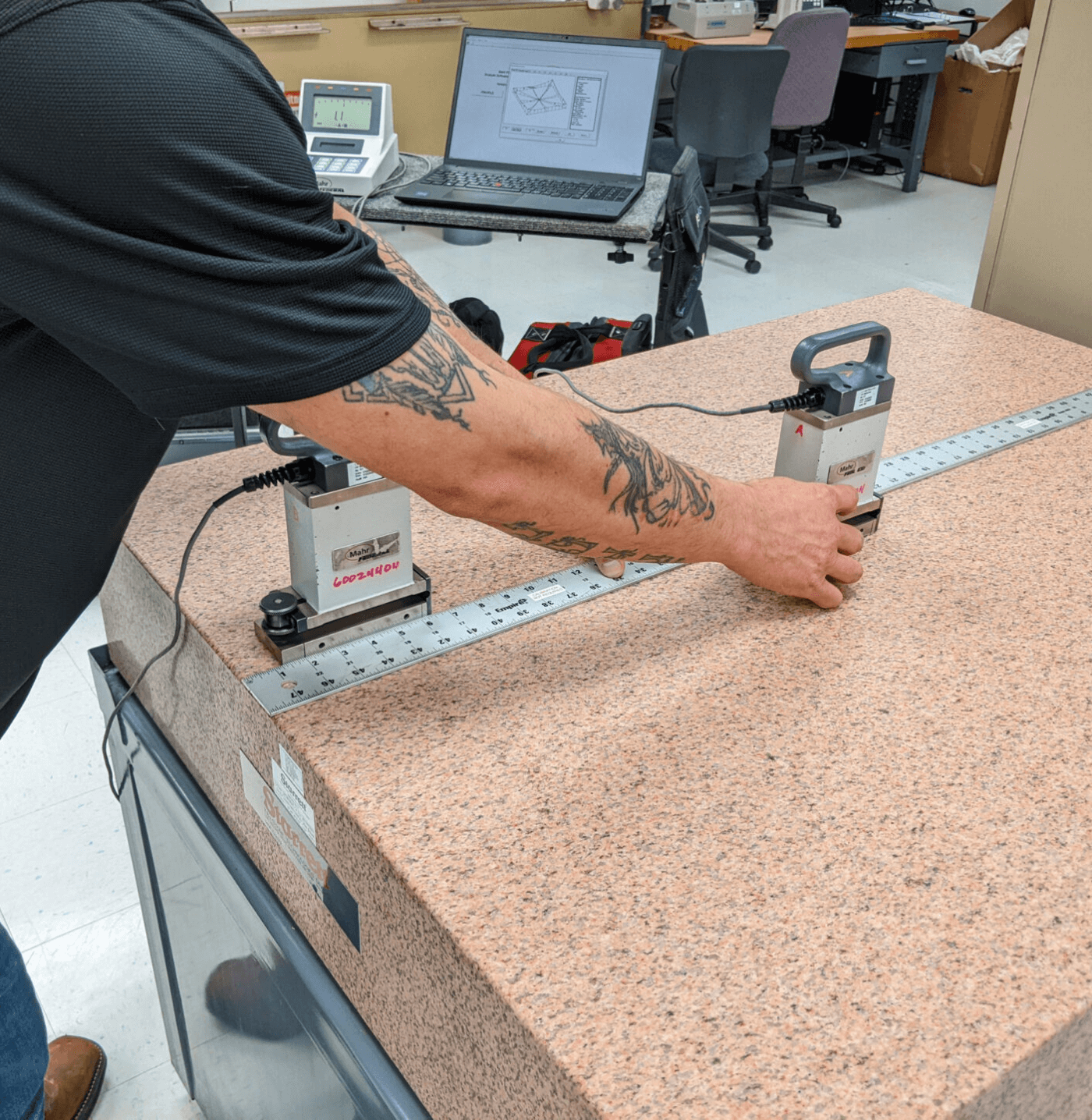 Person with tattoos measuring a large granite surface plate using two precision instruments connected by a metal ruler in a workshop.