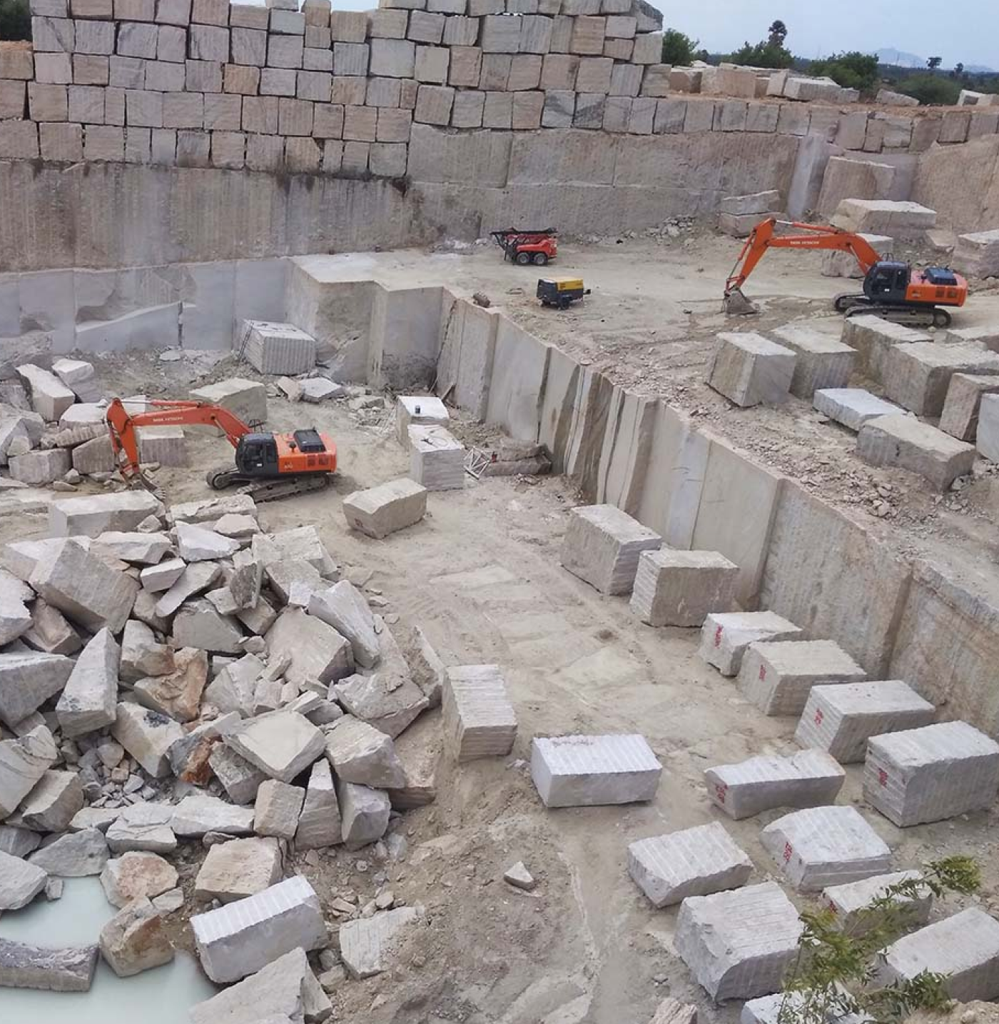 Quarry site with large cut stone blocks, piled rocks, and two orange excavators working on rough terrain.